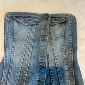 Blå jeanskorsett med knappar - Säljer en snygg blå jeanskorsett med knappar framtill och två fejkfickor upptill. Toppen är axelbandslös och har smockad baksida för extra stretch samt rå kant nertill för en cool look. Perfekt för dig som vill sticka ut med denim.
