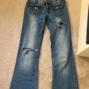 Blå bootcut jeans Gina Tricot 158 - Snygga blå bootcut jeans från Gina Tricot i storlek 158. Jeansen har slitna detaljer och hål på benen, samt två framfickor med lock och knappar. Klassisk femficksmodell i mjukt denimtyg. Egen gjorda hål.