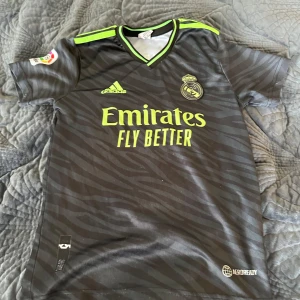 Real Madrid Vinicius Jr. bortatröja Adidas - Snygg Real Madrid bortatröja med Vinicius Jr. 20 på ryggen. Svart med mörkgrå zebramönster och neongula detaljer, Adidas-logga och klubbmärke på bröstet. Tillverkad i lätt funktionsmaterial, perfekt för fotbollsträning eller match.