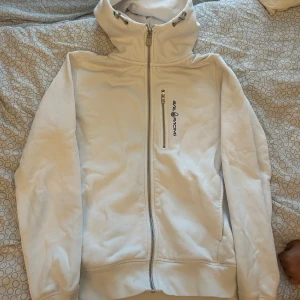 Vit fleecejacka från Sail Racing XS - Snygg vit fleecejacka från Sail Racing med dragkedja framtill och hög krage med snörning. Jackan har broderad logga på bröstet och en praktisk bröstficka med dragkedja. Insidan är mjuk och fluffig, perfekt för kyliga dagar.