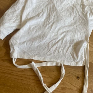 Vit omlottopp med knyt från Nelly - Säljer en vit omlottopp från Nelly i mjuk bomull. Toppen är kortärmad med en öppen rygg där bak och två band som gör att det kan knytas och bli tajt . Perfekt att styla med jeans eller kjol för en fräsch vibe.