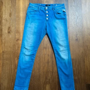 Blå Replay jeans Pilar, storlek 29/28 - Snygga blå jeans från Replay, modell Pilar, med knäppning framtill. Jeansen har en smal passform och är tillverkade i mjukt denimtyg. Fina detaljer med Replay-logga på knappar och ficka. Längd 95 cm, bredd 40 cm. Fraktar oftast samma dag!