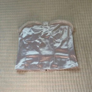 Silver korsettopp från SHEIN XS - Snygg silverfärgad korsettopp från SHEIN i glansigt satinliknande material. Toppen är axelbandslös med spetsdetaljer upptill och har dragkedja bak för enkel påklädning. Perfekt för att sticka ut med en trendig look.