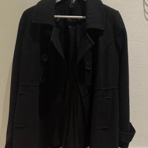 Svart kappa med stora knappar - Stilren svart kappa från Cubus med stora knappar framtill, markerad krage och snygga fickor. Jackan har lång ärm och dekorativa knappar bak i midjan. Perfekt för dig som gillar klassisk och clean look.