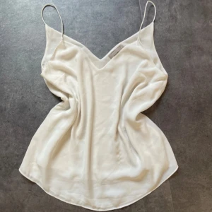 Vit linnetopp med smala band - Supersnygg vit linnetopp i siden med tunna axelband och v-ringning. Toppens material ger en lyxig känsla och den har en lätt glansig finish. Perfekt att styla med jeans eller kjol för en clean look. 