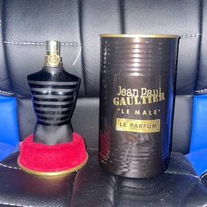 Jean Paul Gaultier Le Male Le Parfum - Snygg parfymflaska i form av en svart manlig torso med matta och blanka ränder. Kommer med originalförpackning i metall med gulddetaljer och röd sammetsbas. Doften är intensiv och maskulin, perfekt för dig som gillar statement-parfymer.