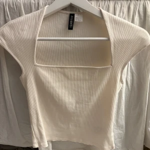 Ribbad vit topp från Divided - Snygg ribbad topp i vitt från Divided med fyrkantig halsringning och ärmlös design. Tunt, stretchigt material som sitter skönt och ger en modern look. Perfekt att styla med jeans eller kjol för en trendig outfit.