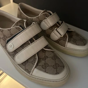 Gucci sneakers med kardborreband - Snygga Gucci sneakers i beige och vitt med klassiskt GG-monogram över hela skon. Skorna har vita detaljer i skinn, kardborreband och diskret logga på sidan. Baksidan har den ikoniska gröna och röda randen. Bra skick. Kontakta för fler bilder. De är självklart äkta och köpta på Vestiaire 