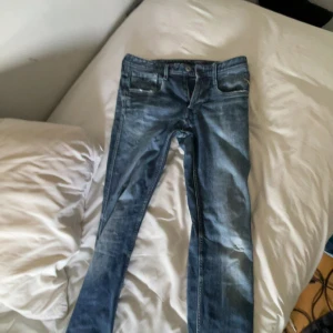 Replay anbass - Säljer ett par klassiska blå jeans med snygga slitningar och tvättade detaljer. Jeansen har fem fickor och normal passform, perfekta till vardagslooken. Materialet är jeans och färgen är en mellanblå ton.