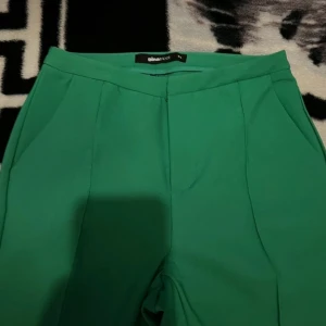 Gröna vida kostymbyxor från Sinaloot - Snygga gröna kostymbyxor från Sinaloot med hög midja och vida ben. Byxorna har klassiska fickor fram och en dekorativ ficka bak. Perfekta för dig som vill sticka ut med färg och stil. Materialet känns mjukt och följsamt.