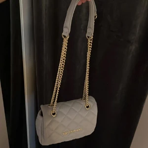 Grå axelväska från Valentino - Snygg beige axelväska från Valentino med quiltad design och guldfärgade kedjor. Väskan har ett lock med magnetstängning och flera fack inuti, perfekt för att hålla ordning på dina grejer. Klassisk och stilren look som lyfter vilken outfit som helst. Inga defekter, precis som ny! Pris går att diskutera ❤️