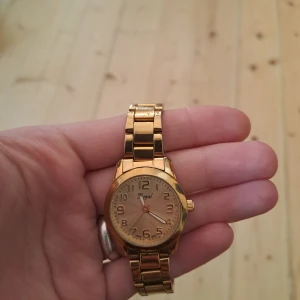 Guldig klocka från Regal - Stilren klocka från Regal i helguldigt utförande med metallarmband och rund urtavla. Klockan har tydliga siffror och klassisk design, perfekt för dig som gillar accessoarer med lite extra bling. Länken är justerbar för att passa handleden.