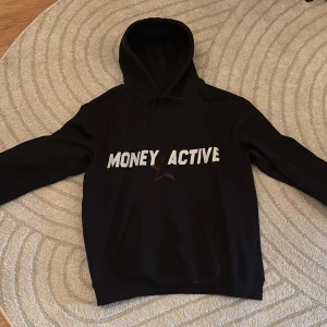 Svart hoodie Money Active - Svart hoodie från Money Active med vit text på bröstet och en liten stjärndetalj. Klassisk modell med huva och dragsko samt känguruficka framtill. Perfekt för dig som gillar streetwear och vill ha en enkel men snygg look.