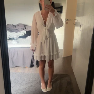 Vit kort omlottklänning med volang - Superfin vit kort klänning med omlottknytning framtill, v-ringning och långa, luftiga ärmar. Klänningen har smock i midjan och volangdetaljer längst ner på kjolen och vid ärmsluten. Perfekt för sommarens alla tillfällen! Från dressy, använd en gång, köpt för 600kr 