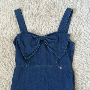 Blå jeansklänning med rosett - Supersöt blå jeansklänning med breda axelband och stor rosett framtill. Klänningen har en rak passform och dragkedja i ryggen. Perfekt för dig som vill ha en chill men ändå snygg look. Liten metallhjärta-detalj på sidan.