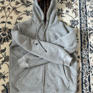 Grå hoodie från Burberry med dragkedja - Snygg grå hoodie från Burberry med klassiskt rutigt foder i huvan. Tröjan har dragkedja, snörning i huvan och broderad logga på bröstet. Tillverkad i mjuk bomullsmix och har två fickor framtill. Perfekt för dig som gillar stilren streetwear.