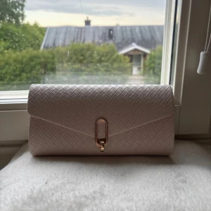 Beige kuvertväska från Anna Field - Snygg beige kuvertväska från Anna Field med flätad struktur och guldfärgad kedja. Väskan har ett elegant spänne framtill och är perfekt när du vill lyfta din outfit med en stilren accessoar. Rymmer det viktigaste och har en lyxig känsla.