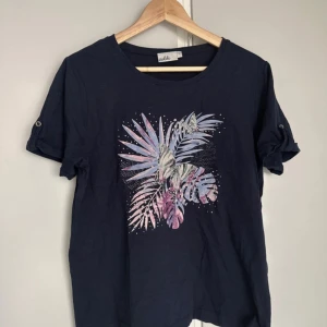 Mörkblå t-shirt med blomtryck Isolde Large - Mörkblå t-shirt från Isolde med korta ärmar och knapp på ärmarna. Framsidan har ett färgglatt blommotiv i lila, rosa och blått med små vita prickar. Skön passform och rund halsringning, perfekt för en chill look.