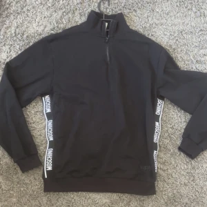 Svart half zip tröja från Moschino - Svart långärmad tröja från Moschino med half zip och hög krage. Snygga vita logoband längs sidorna. Köpt för 2 månader sen och använd 2-3 gånger, pris kan diskuteras. (Den är small men sitter mer som en M/L)