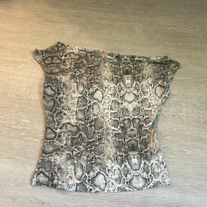 Ormmönstrad offshoulder topp från Nelly - Säljer en snygg offshoulder topp från Nelly med ormmönster i beige, grått och svart. Toppen har en tight passform och är tillverkad i ett mjukt, stretchigt syntetmaterial. Perfekt för dig som vill sticka ut med ett djurmönster och trendig look. Den är lite offshoulder. 