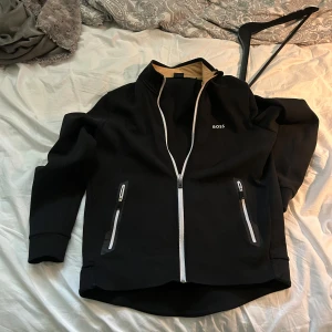 Svart vindjacka från BOSS med dragkedja - Svart tröja  från BOSS med vit dragkedja framtill och på fickorna. Tröjan har hög krage, diskret BOSS-logga på bröstet och är helsvart med stilrena detaljer. Perfekt för dig som gillar en clean och sportig look.