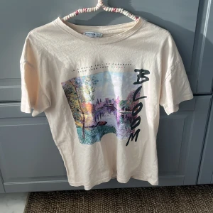 Beige t-shirt med konstmotiv från Stradivarius - Beige t-shirt från Stradivarius med färgstarkt konstmotiv framtill och texten 'BLOOM'. T-shirten har rund halsringning och korta ärmar. Materialet är mjuk bomull och trycket har en konstnärlig vibe med pastelliga färger och stadsmotiv.