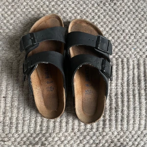 Svarta Birkenstock sandaler i skinn - Klassiska svarta Birkenstock sandaler med två breda remmar i skinn och justerbara spännen. Fotbädden är i kork och sulan är svart med vågigt mönster. Perfekta för sommaren och riktigt sköna att gå i. Nästan nya 9/10 skick