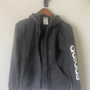 Svart hoodie från Adidas med tryck - Svart hoodie från Adidas med vit logga längs ena ärmen. Klassisk modell med huva, dragkedja och två stora fickor framtill. Perfekt för en sportig och avslappnad stil. Tillverkad i mjuk bomullsblandning för extra komfort.