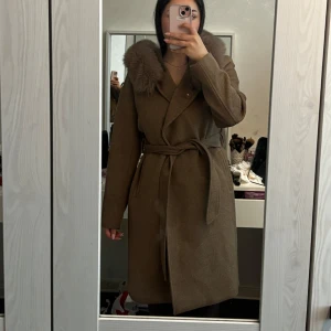 Rockandblue kappa med pälskrage och bälte (modell Talia) - Snygg brun/kamel/ beige kappa med stor fluffig pälskrage och knytskärp i midjan. Jackan har huva, långa ärmar och en rak, klassisk passform. Perfekt för kyliga dagar när du vill vara både varm och stilren. Använd endast en gång. Mycket bra skick. Storlek 38🧥🐻🍂