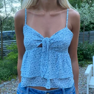 Blommig blå linnetopp med knyt - Supersöt blå linnetopp med små blommor, smala axelband och volangig nederdel. Toppen har en snygg knytning framtill och en liten cut-out under bysten. Perfekt för varma dagar och ger en fräsch sommarkänsla.