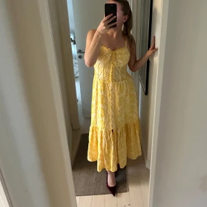 Gul långklänning med broderade detaljer - Superfin gul långklänning med broderade mönster och volang nertill. Klänningen har smala axelband, söt rosett framtill och en luftig passform. Perfekt för varma dagar och ger en riktigt somrig vibe.