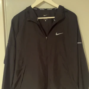 Svart Nike vindjacka  - Svart vindjacka från Nike med huva och dragkedja framtill. Jackan har diskreta reflexdetaljer på ärmarna och klassisk Nike-logga på bröstet. Perfekt för löprundan eller när du vill ha en sportig look. Lätt och smidig modell med fickor på sidorna.