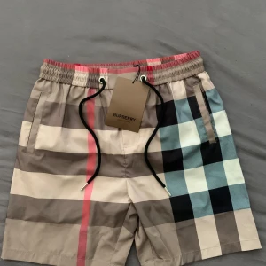 Burberry rutiga shorts med snörning - Snygga Burberry shorts med klassiskt rutigt mönster i beige, svart, blått och rött. De har elastisk midja med svart snörning och fickor på sidorna samt bak. Perfekta för sommaren och ger en lyxig vibe till din outfit. Helt ny!