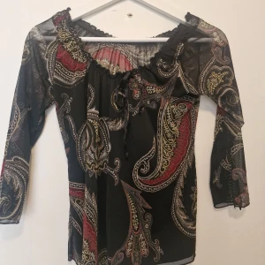 Svart mesh blus med paisleymönster - Säljer en svart meshblus med paisleyinspirerat mönster i rött, beige och gult. Blusen har trekvartsärmar, lätt transparent tyg och en söt knytning vid halsen. Perfekt för dig som gillar boho-vibbar och unika detaljer.