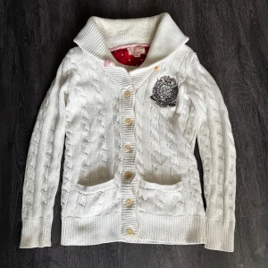 Vit kabelstickad kofta Polo Jeans - Vit kabelstickad kofta från Polo Jeans Company med bred krage, knappar framtill och två fickor. Snygg broderad logga på bröstet och ribbade muddar. Perfekt för lager-på-lager och chill höststil.