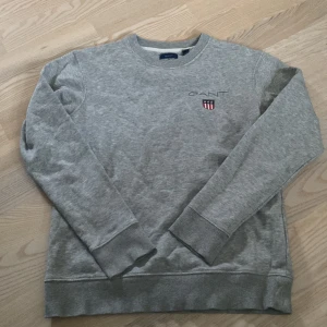 Grå sweatshirt från GANT - Klassisk grå sweatshirt från GANT med broderad logga och sköld på bröstet. Tröjan har rund hals, ribbade muddar och är tillverkad i mjuk bomull. Perfekt för en chill och stilren look.