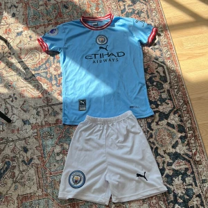 Manchester City Haaland matchställ Puma 152 - Manchester City matchställ med Haaland 9 på ryggen. Tröjan är ljusblå med röda och vita detaljer vid krage och ärmslut, kortärmad och tillverkad i funktionsmaterial. Byxorna är vita med klubbmärke och Puma-logga. Perfekt för fotbollsfans!