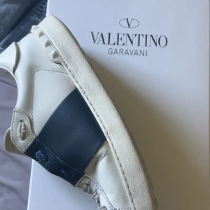 Valentino Garavani Open sneakers - Snygga Valentino Garavani Open sneakers i vitt skinn med en bred marinblå läderdetalj över sidan. Klassisk platt sula och ikoniska nitar baktill. Perfekta för dig som gillar exklusiva och stilrena sneakers. ‼️Intressekoll‼️har box påse, dustbag. O ett kuvert. Skicka pris förslag! Stolek 39 men passar 40!