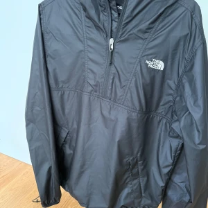 Svart vindjacka från The North Face - Svart vindjacka från The North Face med huva och halv dragkedja. Jackan har två fickor framtill och justerbar dragsko nertill. Perfekt för blåsiga dagar och enkel. Snygg och stilren design med logga på bröstet. Knappt använd så är i ypperligt skick. Online orderbekräftelse finns, hör av er vid frågor och funderingar. Mvh!