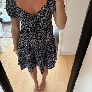 Blommig kortklänning med puffärm - Supersöt marinblå kortklänning med vita små blommor, puffärmar och v-ringning med knyt framtill. Klänningen har en luftig passform och volang i kjolen. Aldrig använd