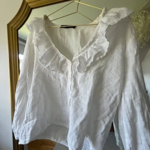 Vit volangblus i linne från Zara - Supersöt vit blus från Zara i luftig linnemix. Skjortan har v-ringning med volangdetaljer längs kragen och knappar framtill. 💕