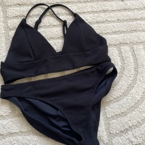 Svart ribbad bikini med trekantsöverdel - Snygg svart bikini med ribbad struktur och trekantsformad överdel med smala axelband. Underdelen har klassisk passform och materialet känns mjukt och stretchigt. Perfekt för strandhäng eller poolparty.