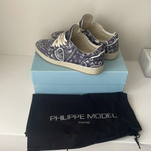 Philippe Model blå sneakers bandanamönster - Snygga sneakers från Philippe Model i marinblått med vitt bandanamönster. Skorna har vita snören, platt sula och rund tå. Tillverkade i skinn med klassisk Philippe Model-logga på sidan. Perfekta för dig som vill sticka ut med en cool och unik look. Priset är inte hugget i sten 😊