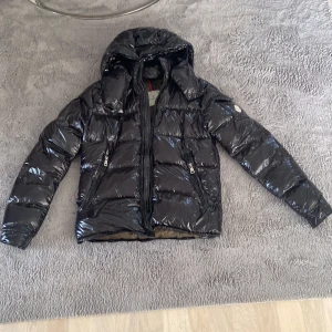 Svart Moncler pufferjacka med huva - Säljer en glansig svart pufferjacka från Moncler med huva och dragkedja. Jackan har två snedställda fickor med dragkedja framtill och en extra ficka på ärmen med Moncler-logga. Insidan är olivgrön med en cool serietidningspatch. Perfekt för kalla dagar. Det ända problemet är att dragkedjan inte fungerar.