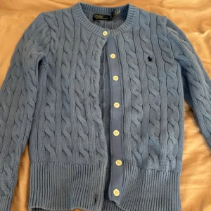 Blå kabelstickad kofta från Polo Ralph Lauren - Kabelstickad blå kofta från Polo Ralph Lauren med rund halsringning och vita knappar framtill. Klassisk broderad logga på bröstet och ribbade muddar. Perfekt för lager-på-lager och snygg till jeans eller kjol.