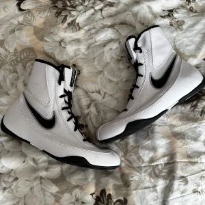 Nike Machomai boxningsskor vit/svart - Nike Machomai boxningsskor i vitt med svarta detaljer och svart Nike-logga. Skorna har hög modell med snörning och platt sula för bra grepp. Ovandelen är i syntetmaterial och mesh för lätthet och ventilation. Perfekta för dig som vill ha stilrena och funktionella boxningsskor. Nypris 1300kr