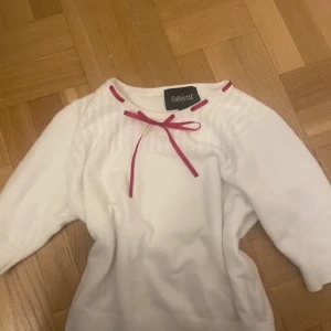 Vit stickad topp med rosett Collectif - Supersöt vit stickad topp från Collectif med trekvartsärm och dekorativ röd rosett vid halsen. Fint hålmönster upptill och figurnära passform. Perfekt för dig som gillar retrovibbar och unika detaljer. Är storlek L men sitter som en S. 