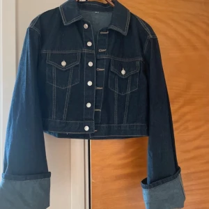 Mörkblå croppad jeansjacka - Snygg mörkblå croppad jeansjacka med klassisk krage, silverfärgade knappar och två bröstfickor med lock. Jackan har långa ärmar med breda uppvik och kontrastsömmar som ger en cool look. 