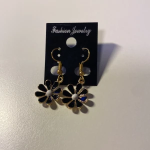Handgjorda Blommiga örhängen i svart och guld *Endast 1 Kvar* - Snygga örhängen med blommor i svart och guld, med vit detalj i mitten. Hängande modell med guldfärgade krokar. Perfekta för att ge din look en cool och lekfull touch.