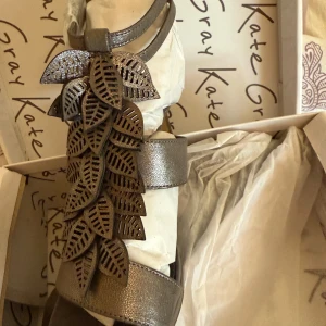 Metallic sandaler med bladdekor Kate Gray - Snygga metallicfärgade sandaler från Kate Gray med öppen tå, hög klack och coola utskurna blad i skinn framtill. Remmar runt vristen och foten ger en trendig look. Perfekta för dig som vill sticka ut med detaljer och glans.